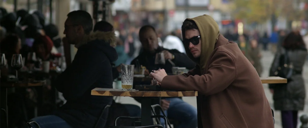 Marked Men: Rule + Shaw (2025) movie still: medium — A bustling sidewalk cafe features several people seated at tables near a city street. A man in…