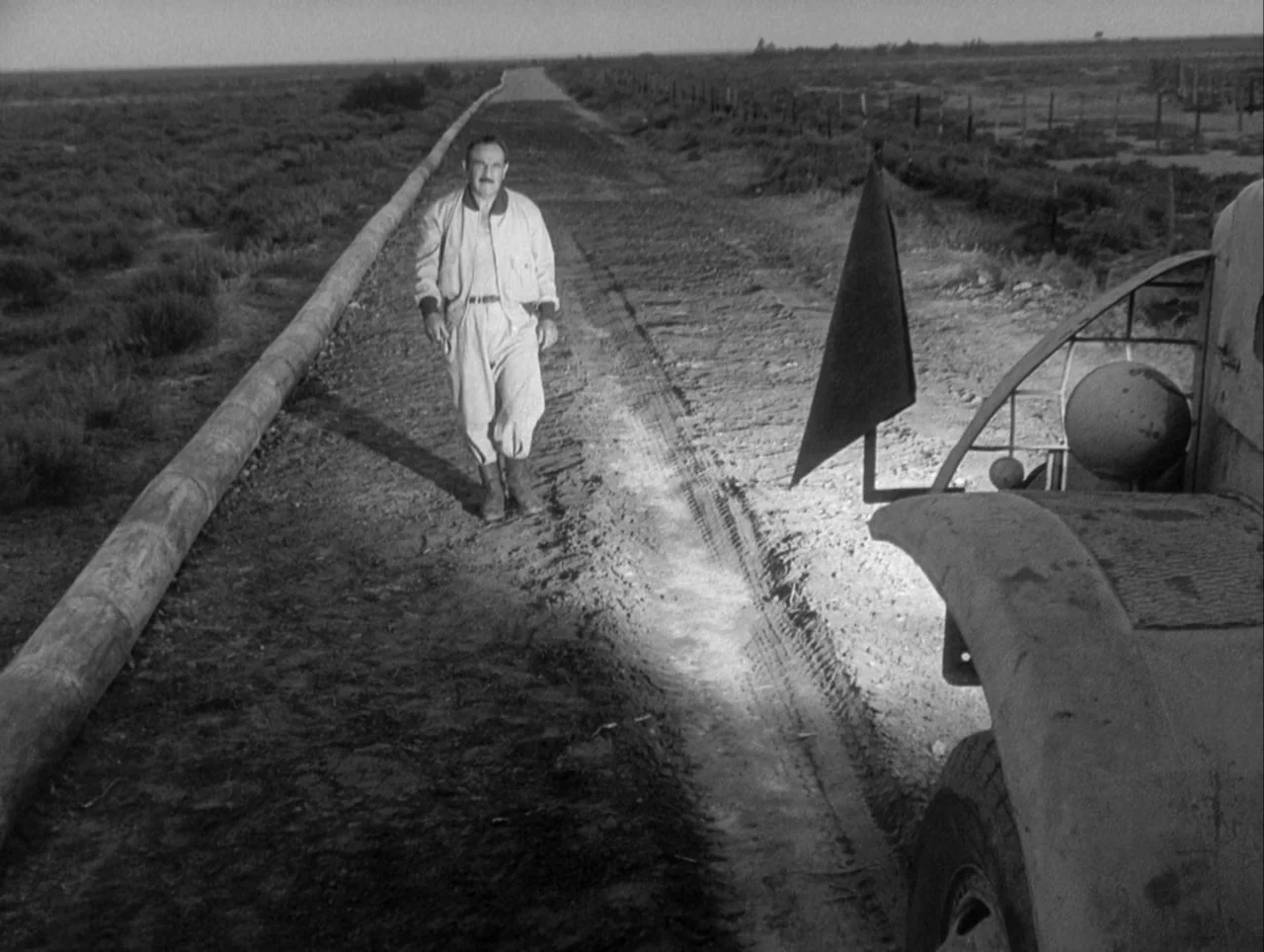 The Wages of Fear (1953) movie still: fullbody — A flat, barren landscape stretches out along a dirt track next to a pipeline. A middle-aged man…
