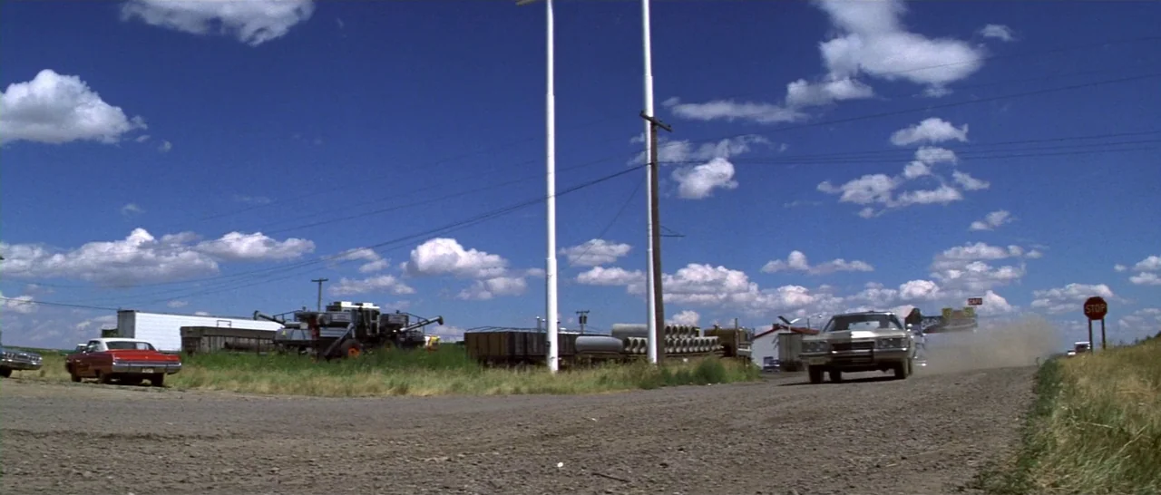 Thunderbolt & Lightfoot (1974) movie still: wide — A rural gravel road under a bright blue sky with large scattered clouds. A vintage car drives…