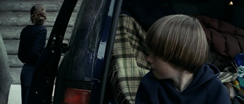 Time Of The Wolf (2003) movie still: medium — A child sits inside the open trunk of a blue SUV filled with blankets. A woman walks away towards a…