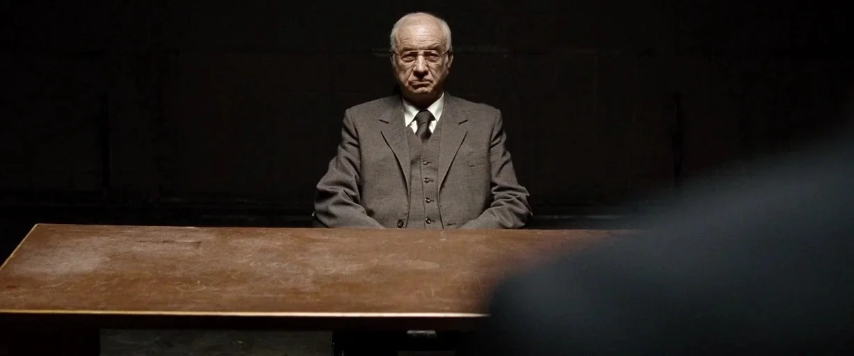 The International (2009) movie still: medium — An older man sits at a wooden table in a dark, empty, windowless space. He is being observed by…