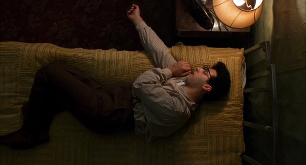 Barton Fink (1991) movie still: medium — A man lies on a single bed with a textured yellow quilt in a dim room. The overhead camera angle looks down…