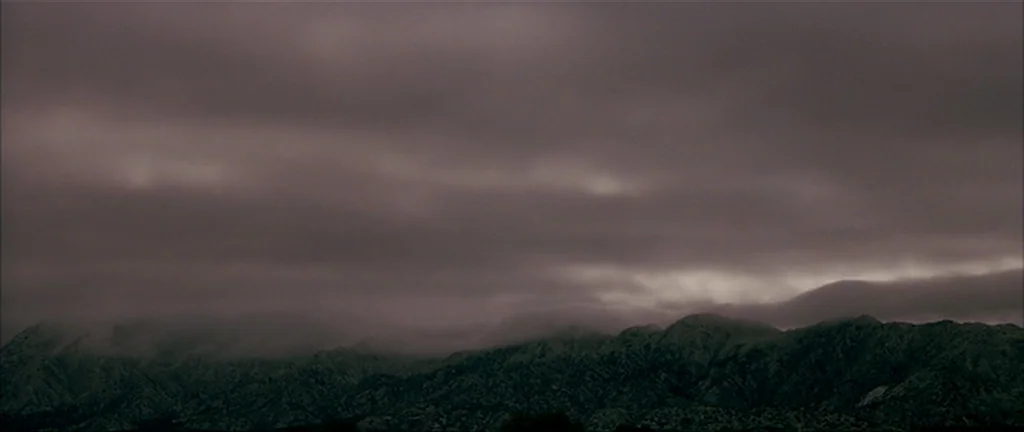 Gerry (2002) movie still: establishing — A rugged mountain range stretches across the horizon under a heavy, dense cloud layer. Dark fog hugs the…