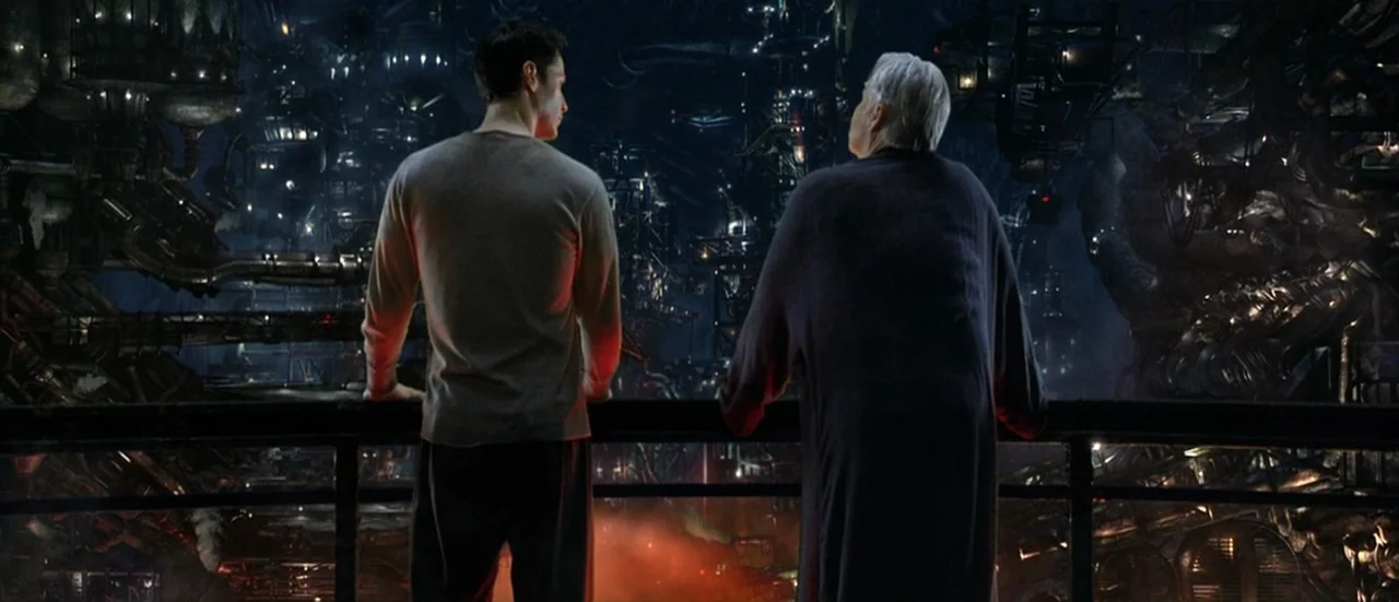 The Matrix Reloaded (2003) movie still: fullbody — A dark subterranean observation deck overlooks a vast and complex industrial mechanical city. Two…