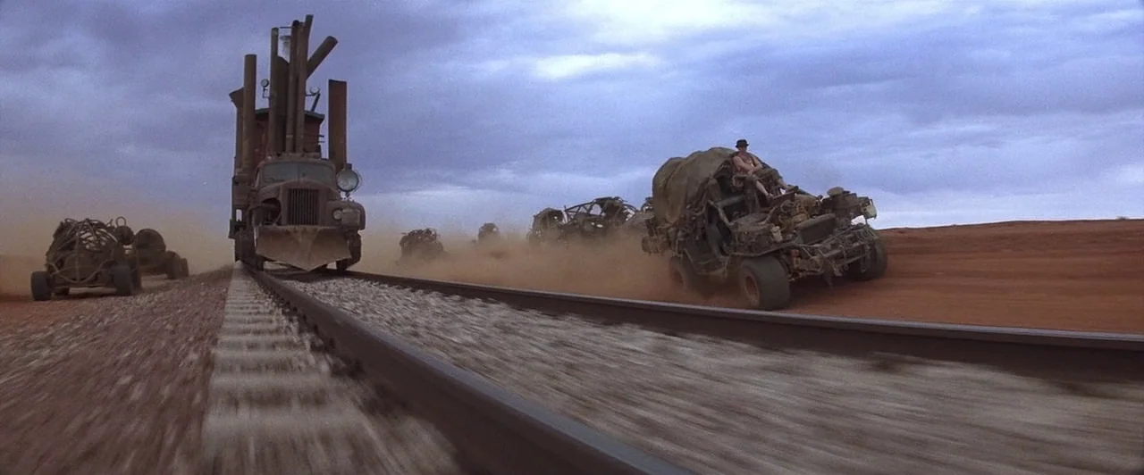Mad Max Beyond Thunderdome (1985) movie still: wide — A group of armored vehicles travels through a vast, dusty, reddish desert landscape. The train…