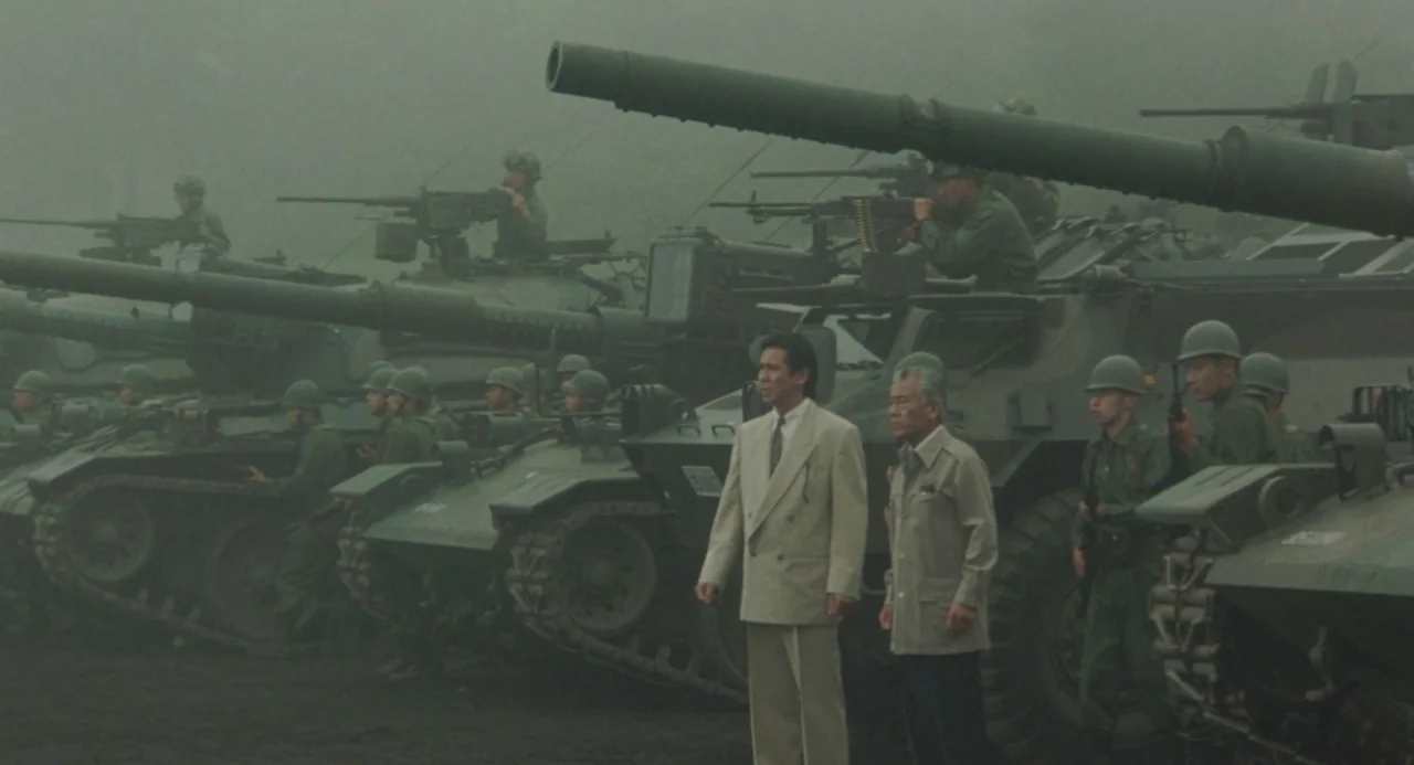 Godzilla vs. King Ghidorah (1991) movie still: wide — A line of armored combat tanks sits on a dark, mist-covered field. Two men in business attire…