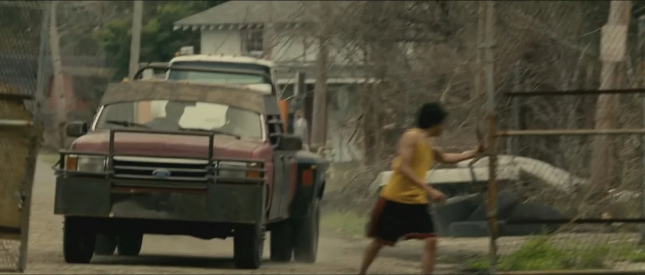 Contraband (2012) movie still: medium — A dusty dirt road with a large red truck and a chain link fence. A young man runs along the roadside fence.