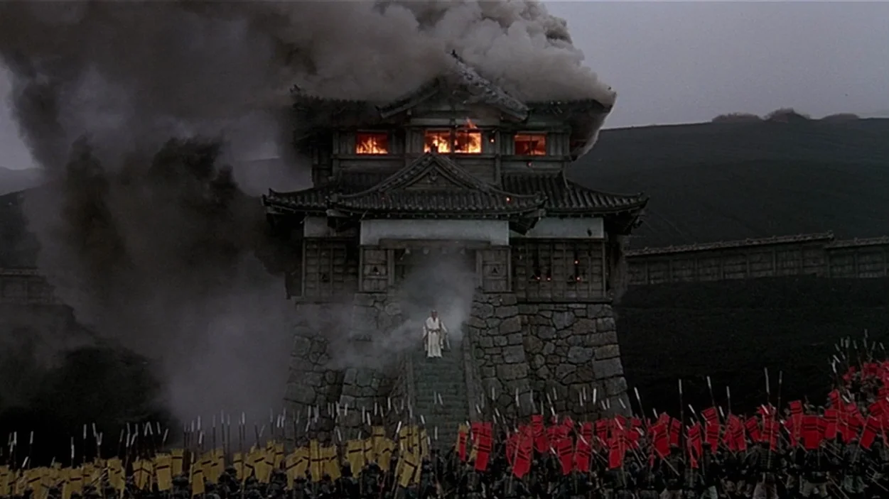 Ran (1985) movie still: wide — A traditional Japanese stone fortress is engulfed in thick smoke and flames from within. A lone figure stands at the…