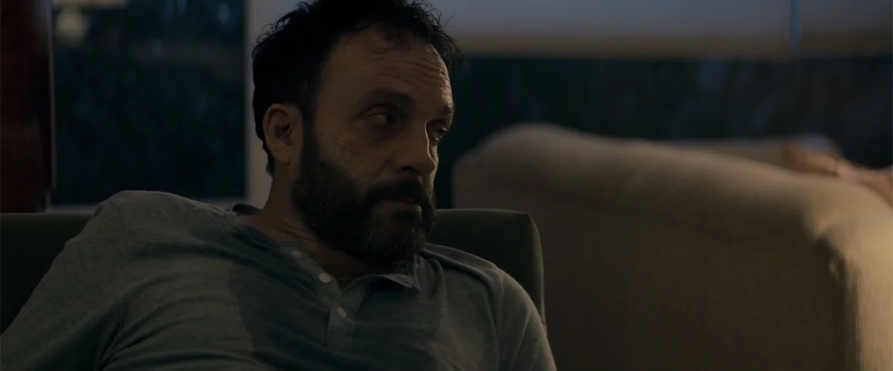 Human Animals (2020) movie still: medium — A dimly lit room interior features a person sitting on furniture during the night. The subject is a…