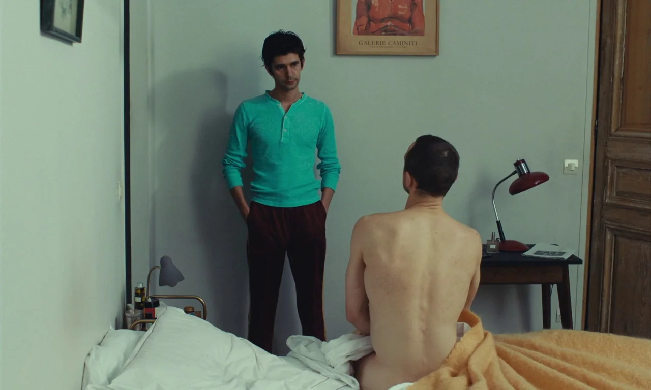 Passages (2023) movie still: fullbody — A minimalist bedroom features light walls adorned with a single framed poster. One man wearing casual attire…