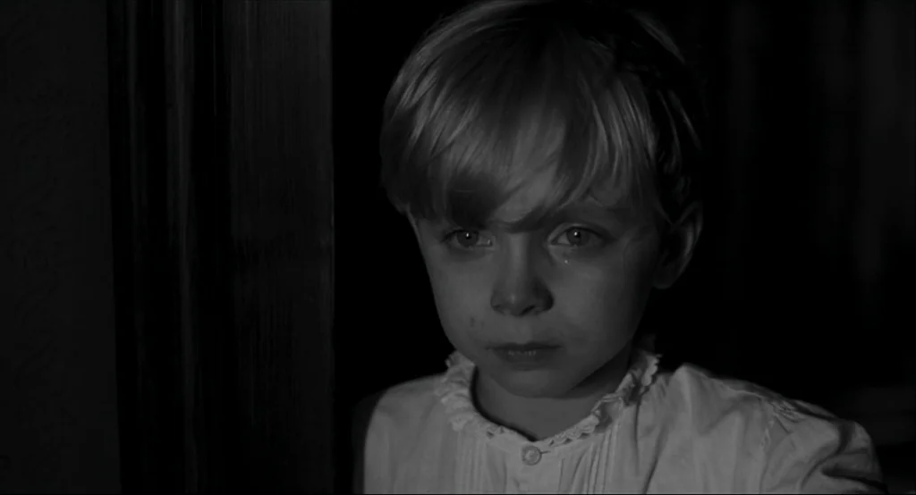 The White Ribbon (2009) movie still: closeup — A dimly lit indoor space with a dark doorway frame on the left side. A young blonde boy stands…