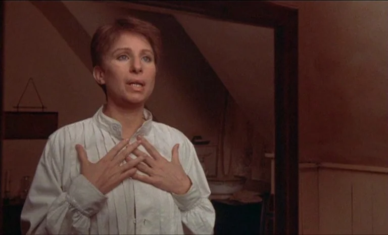 Yentl (1983) movie still: medium — A dimly lit room with sloped attic walls features a woman standing before a mirror. The woman wears a white shirt…