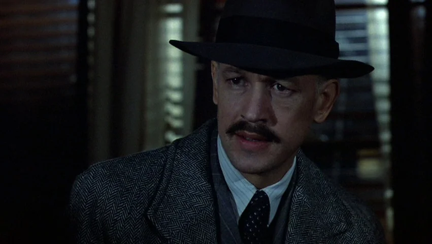 Hammett (1982) movie still: closeup — A dimly lit room features closed wooden blinds casting lines of darkness behind the subject. A man in a wool…