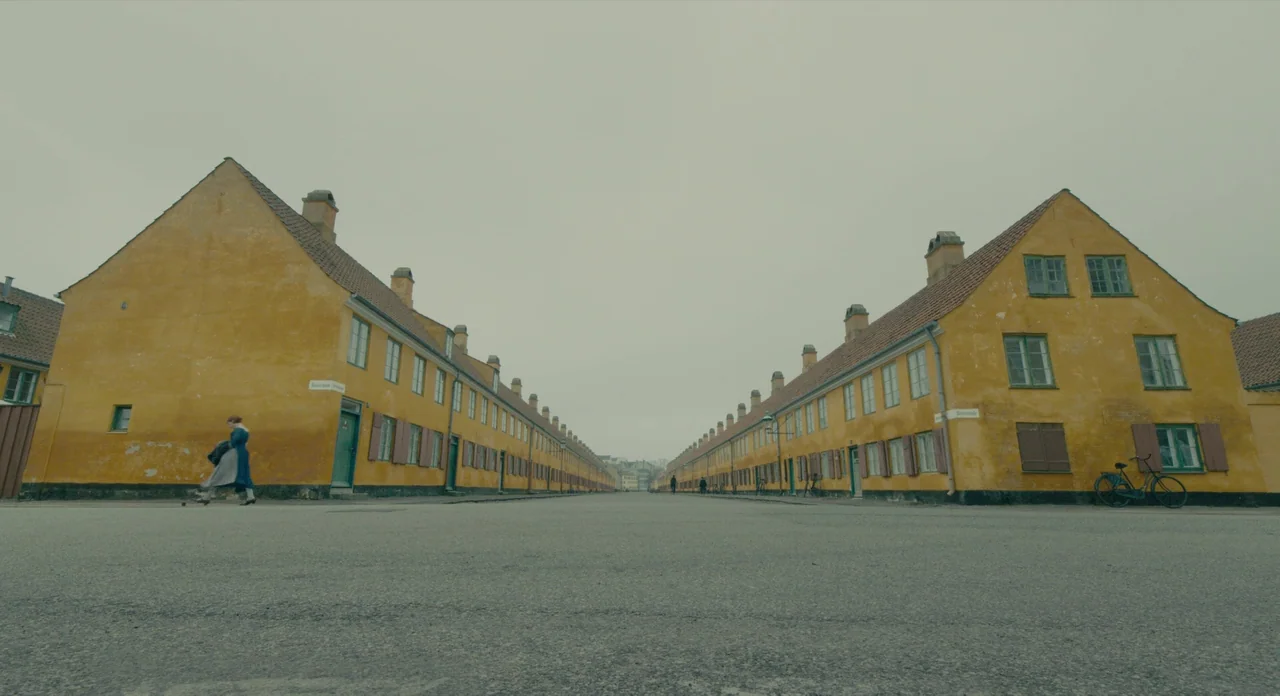 The Danish Girl (2015) movie still: establishing — A symmetrical street is lined with uniform yellow row houses on both sides. A woman walks along…