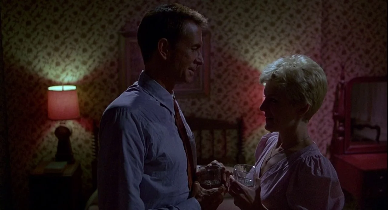 Psycho III (1986) movie still: medium — A dimly lit room features vintage wallpaper with a floral pattern and wooden furniture. A man and a woman…