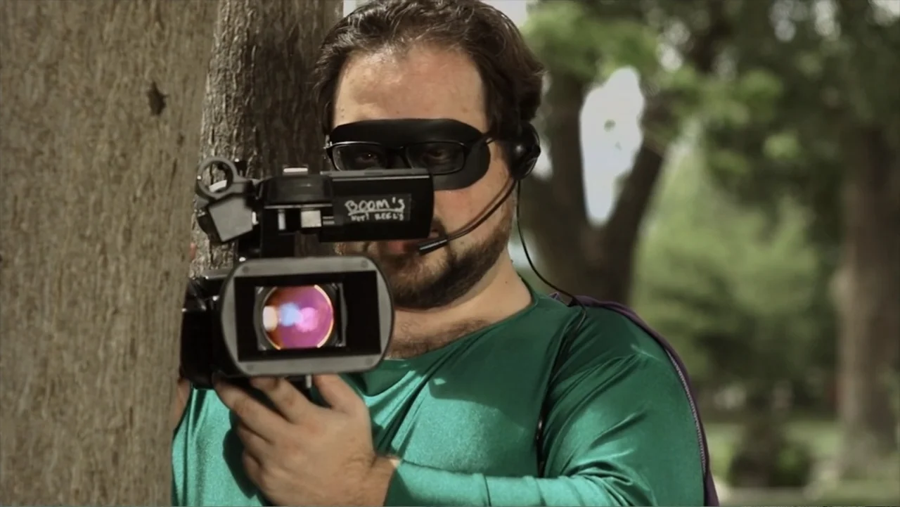 Hello I Love You (2018) movie still: medium — A man in a green costume stands behind a tree in a park. He holds a camcorder while looking directly…