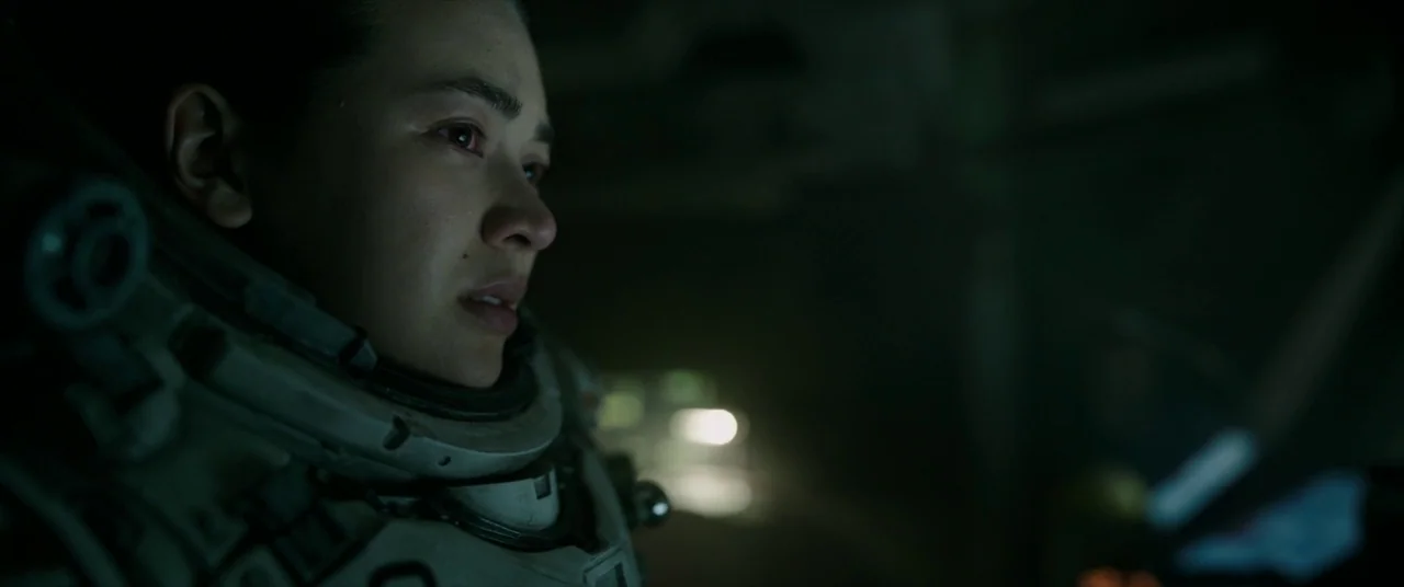 Underwater (2020) movie still: closeup — The interior of a dark, cramped cockpit illuminated by dim monitor lights. A person wearing a space suit…