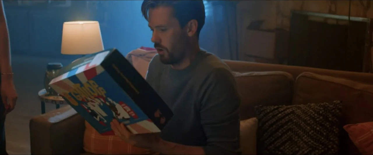 Gatlopp: Hell of a Game (2022) movie still: medium — A living room interior with a sofa and a lit lamp on a side table. A man in a grey sweater sits…