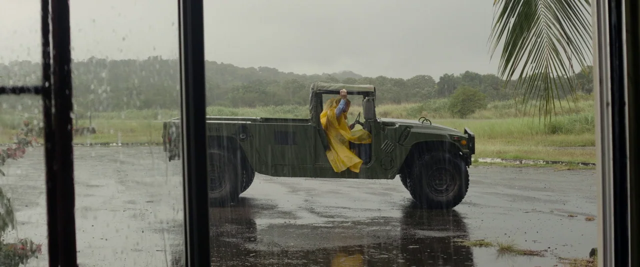 Stars At Noon (2022) movie still: wide — An olive green military humvee sits in a paved lot during a light rain. A man wearing a bright yellow…