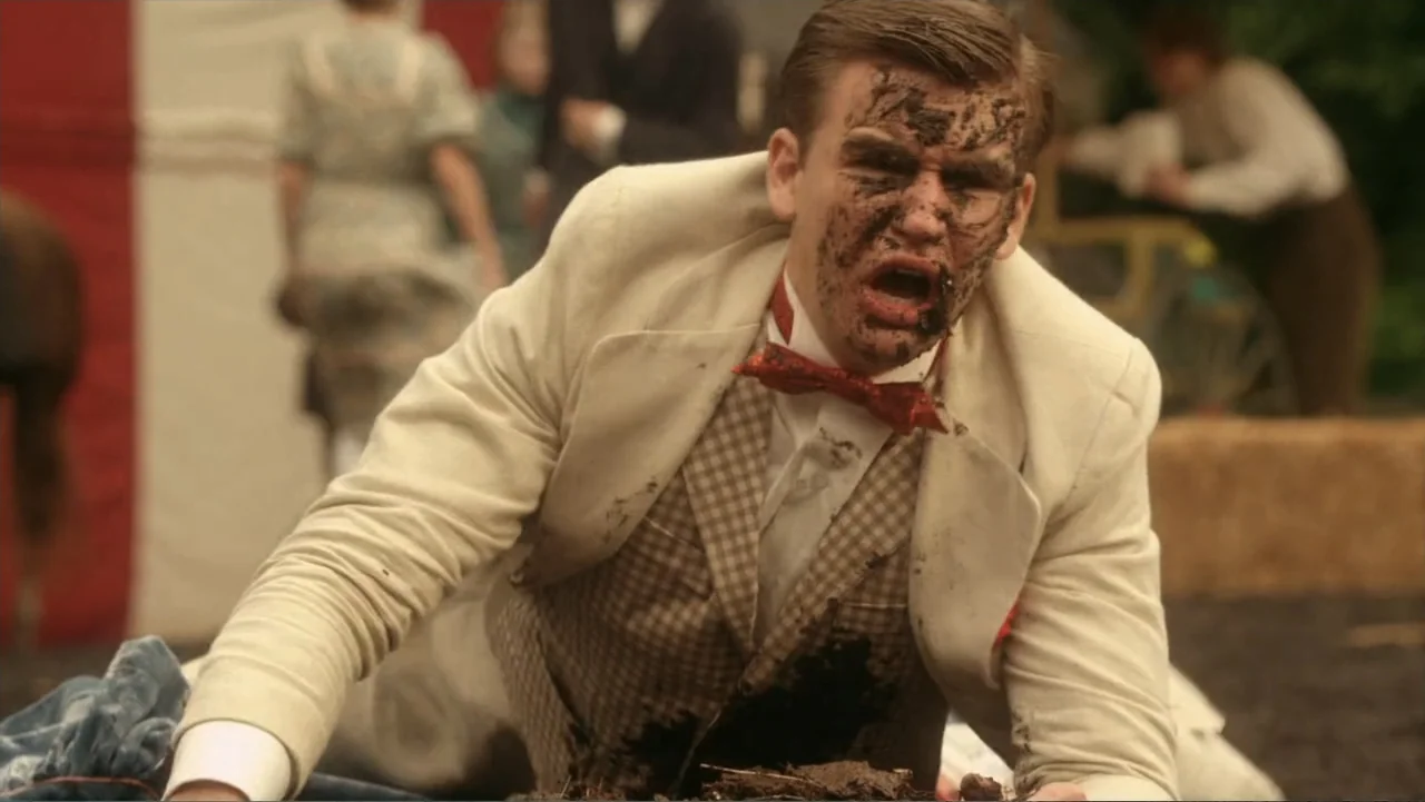 Jinxed (2013) movie still: medium — An outdoor fairground setting with a blurred tent and other figures in the background. A man in a stained white…