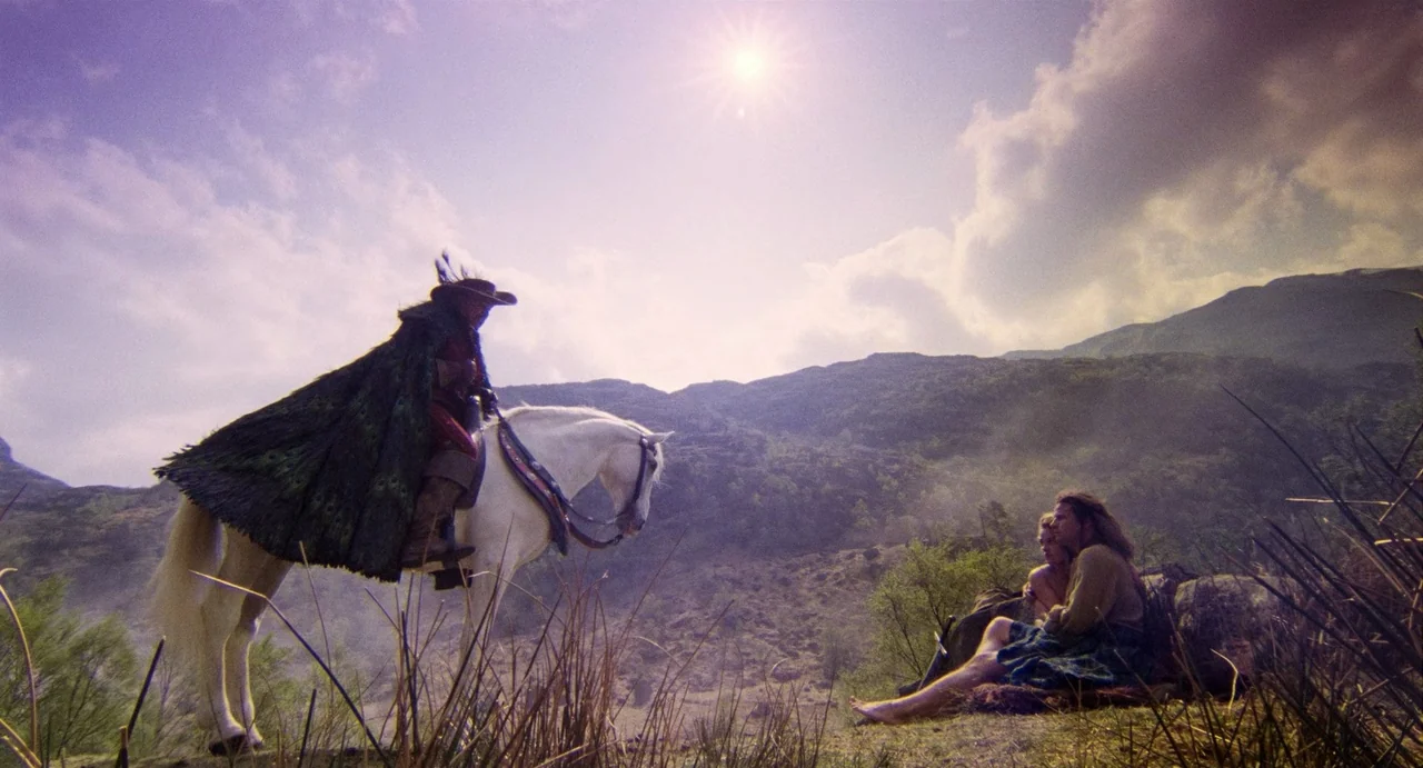 Highlander (1986) movie still: wide — A rugged mountain hillside with tall grasses under a bright sunlit sky. A horseback rider encounters a couple…