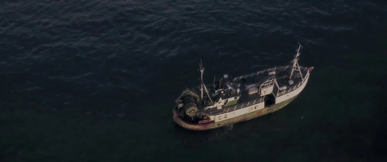 Sea Fever (2019) movie still: wide — A small white and green fishing trawler floats in the middle of dark water. The vessel is viewed from directly…