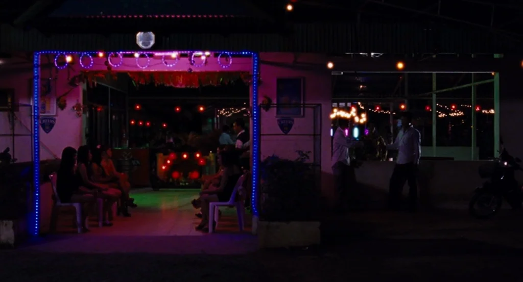 Almayer’s Folly (2011) movie still: wide — An open-air bar at night features bright blue neon lights framing the primary entrance area. Several…
