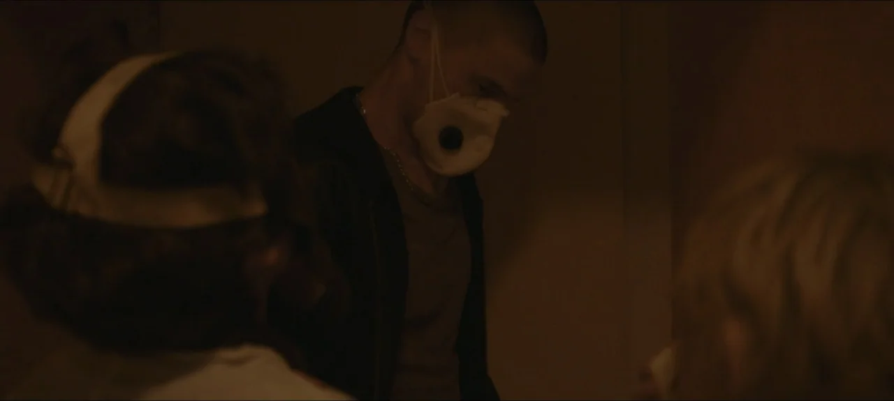 Containment (2015) movie still: medium — A dimly lit room provides a backdrop for two individuals in close proximity. A man wearing a white…
