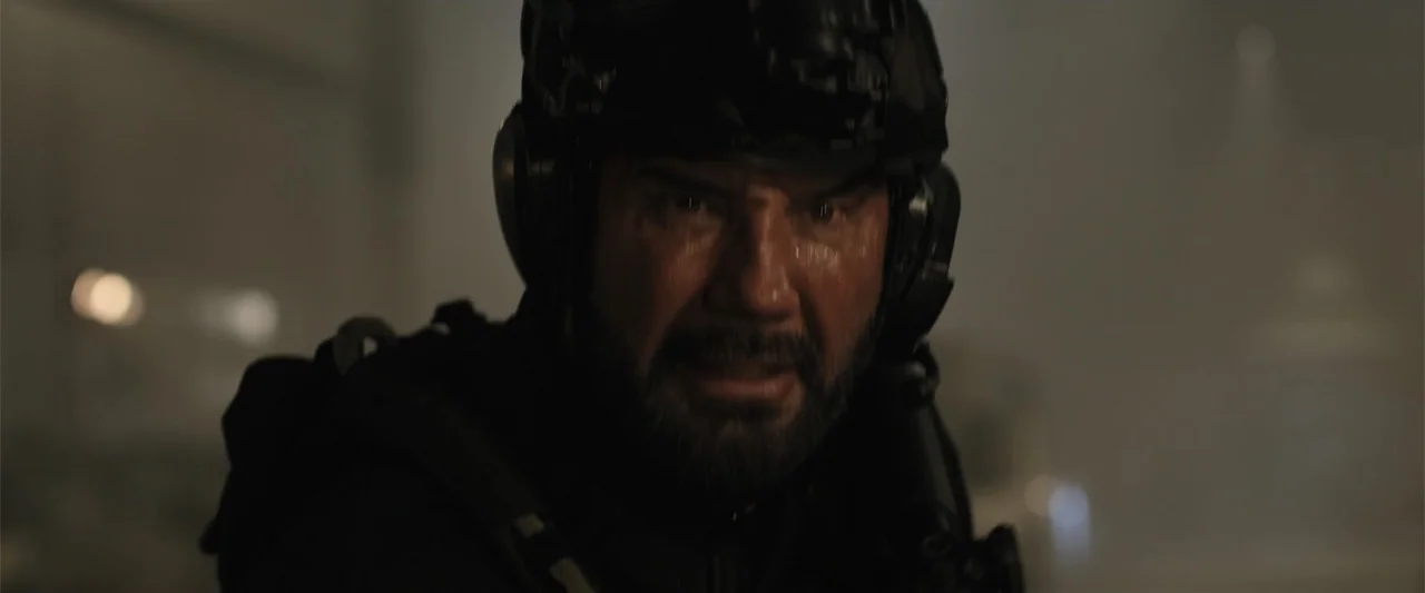 Trap House (2025) movie still: closeup — A dark, cramped space with ambient light suggesting a tactical military environment. A man in tactical gear…