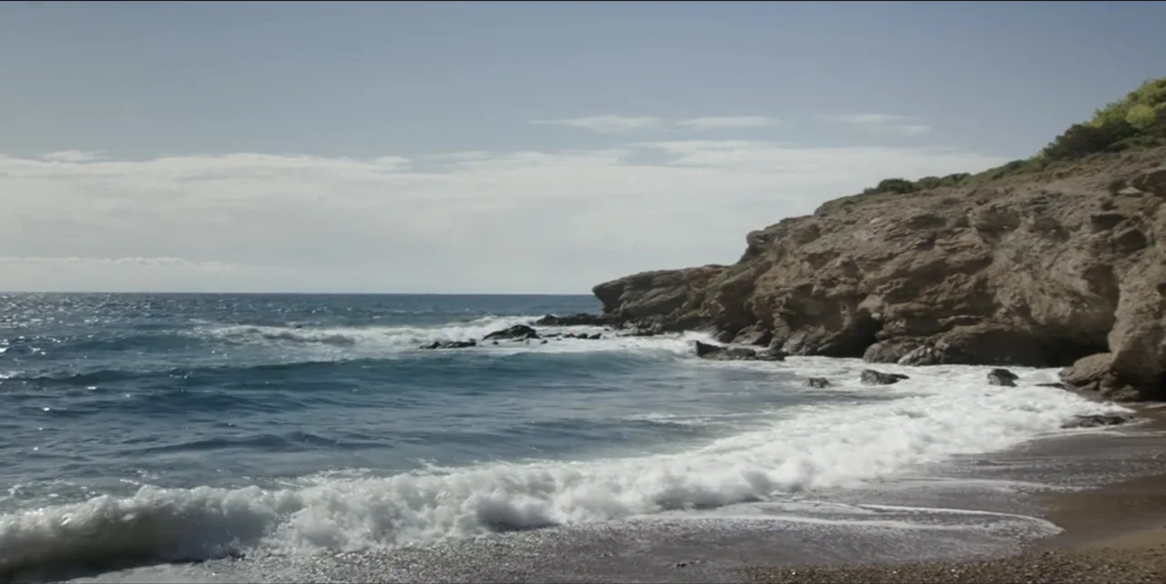 The Hunted (2024) movie still: establishing — A rugged coastline features a dark pebbled beach meeting calm, rolling coastal waves. A rocky cliff…