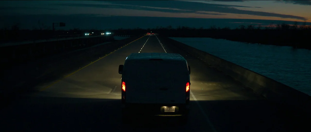 The Killer (2023) movie still: wide — A white cargo van drives along an elevated highway over dark water at night. The surrounding landscape is…