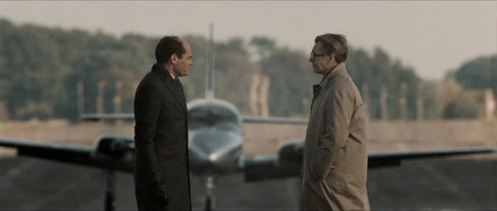 Tinker Tailor Soldier Spy (2011) movie still: fullbody — Two men stand facing each other on an asphalt runway with a small plane behind. Trees and…