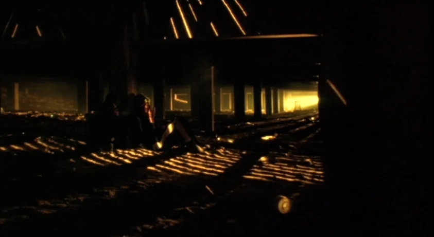 Requiem For A Dream (2000) movie still: wide — The space beneath a pier features repeating wooden support columns and sand-covered ground. A single…