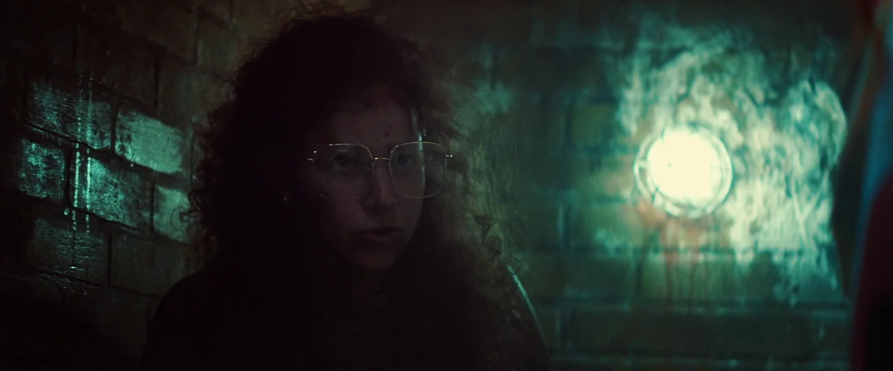 Revealer (2022) movie still: closeup — A dark brick wall provides the backdrop for a dimly lit underground space. A young woman with curly hair…