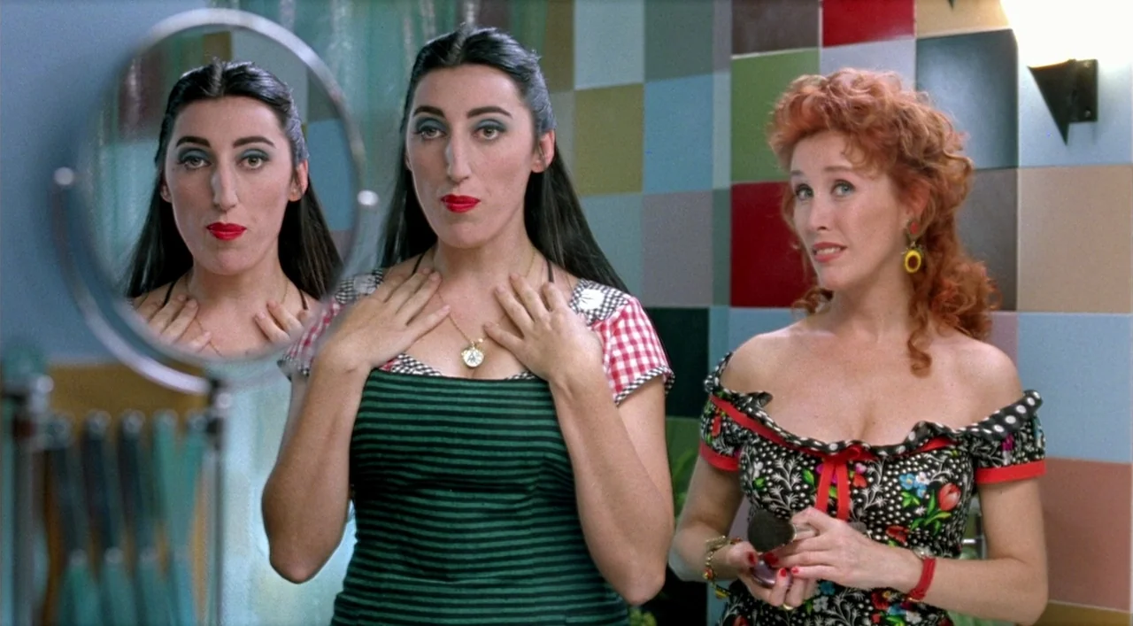 Kika (1993) movie still: medium — A bathroom with a grid of multicolored square tiles on the wall behind two women. The woman on the left adjusts…
