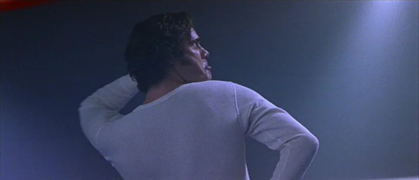 Man On The Moon (1999) movie still: medium — A dark, undefined interior space serves as the setting for the frame. A man in a white long-sleeved…