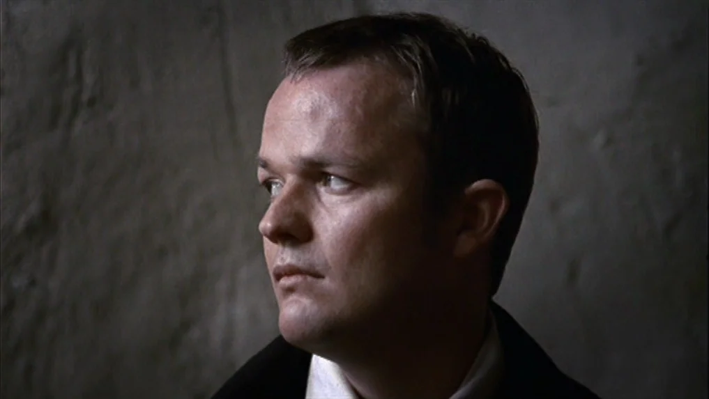 The Scarlet Letter (1973) movie still: closeup — A man stands against a rough, grey wall with a textured stone-like surface. The man gazes to the…