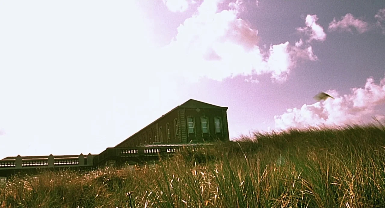 The Diving Bell and the Butterfly (2007) movie still: wide — A long, dark brick building sits on a rise above sand dunes with grass. Tall blades of…