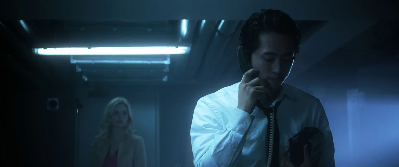 Mayhem (2017) movie still: medium — A dimly lit narrow hallway with cool blue lighting cast by an overhead fixture. A man in a white dress shirt…