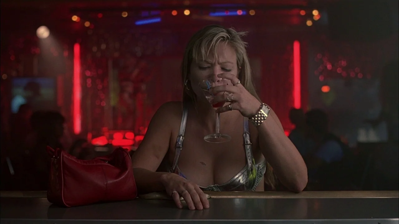 Whore (1991) movie still: medium — A dimly lit nightlife setting with red neon accents blurred in the background. A woman sits at a counter drinking…
