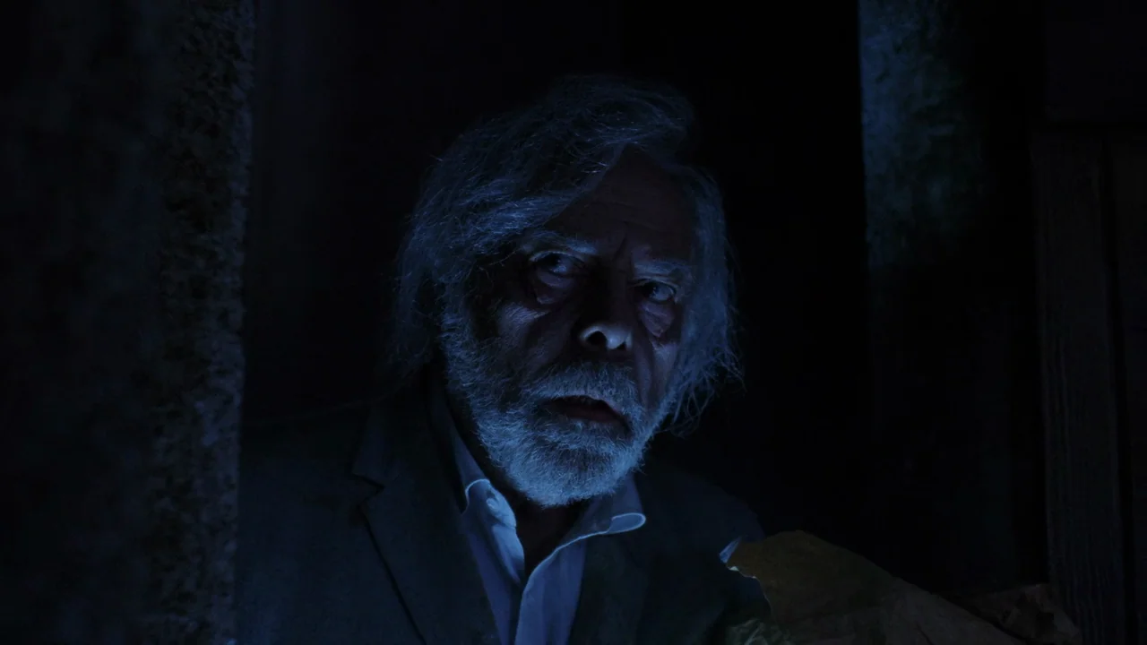 The Sect (1991) movie still: closeup — An elderly man is framed closely in a dark, dimly lit interior environment. The subject looks toward the…