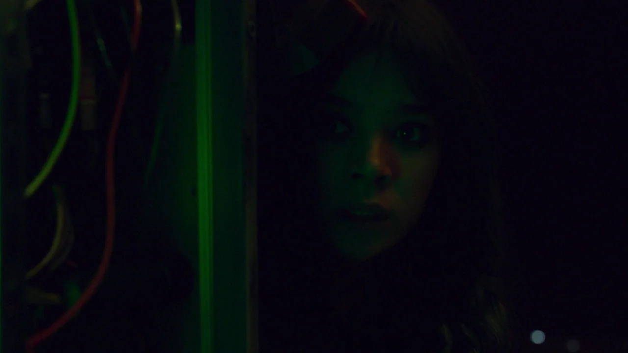 Bumblebee (2018) movie still: closeup — A dark, cramped space features exposed electrical wiring mounted on a green metal frame. A young woman peers…