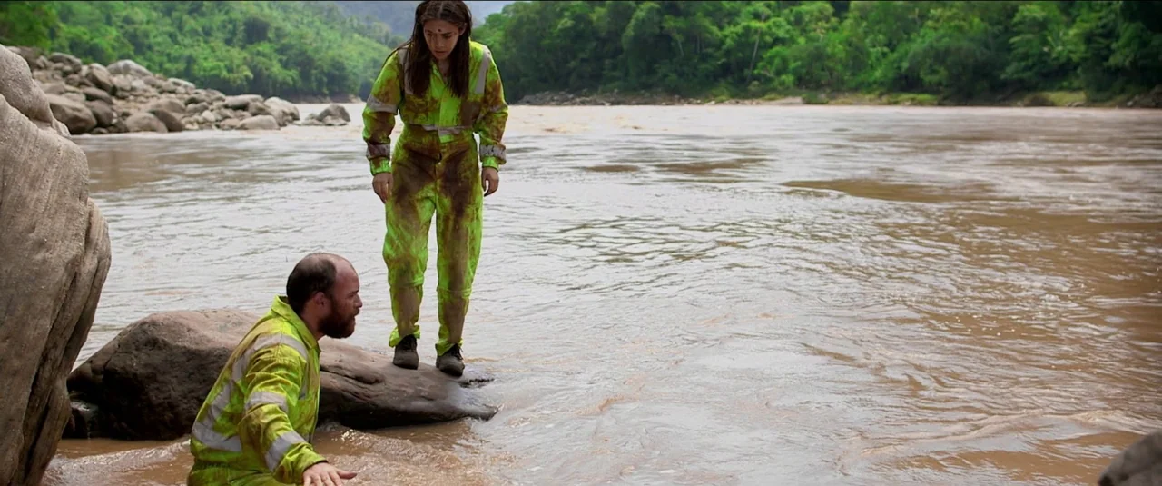 Green Inferno (2013) movie still: wide — A wide, muddy river flows past a rocky bank surrounded by lush green foliage. A man kneels in the shallow…