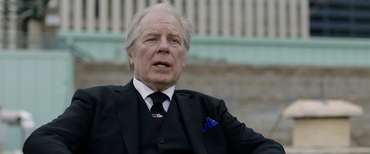 Playing God (2021) movie still: medium — An outdoor rooftop area features a concrete wall and tiered seating in the background. An elderly man in a…