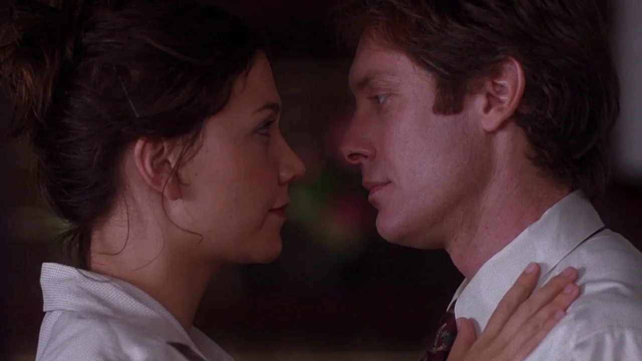 Secretary movie still: medium — A dark indoor office space provides a backdrop for the close interaction of two people. A man and a woman stand face…