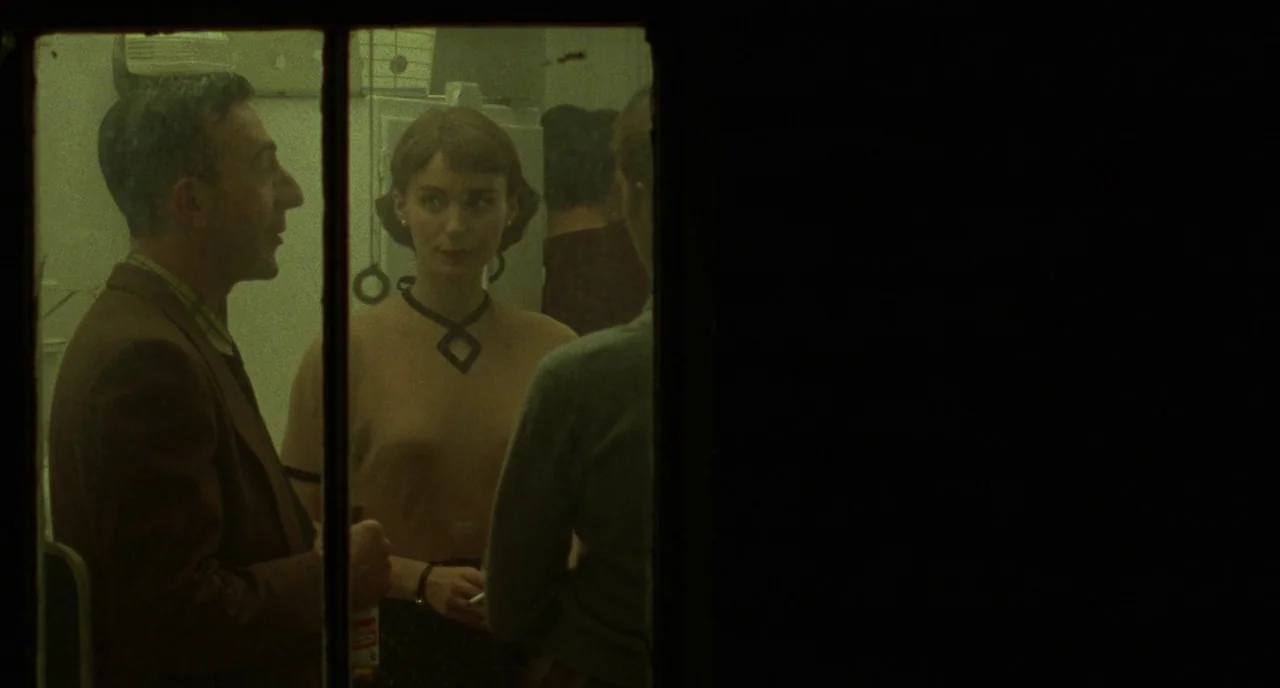 Carol (2015) movie still: medium — A dimly lit indoor space with dark walls viewed through a glass pane. A man and a woman in 1950s attire stand…