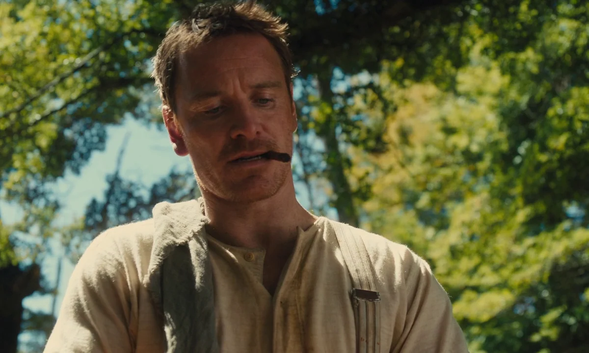 Slow West (2015) movie still: medium — A man stands outdoors with blurry green tree foliage in the background. He looks down while holding a cigar…