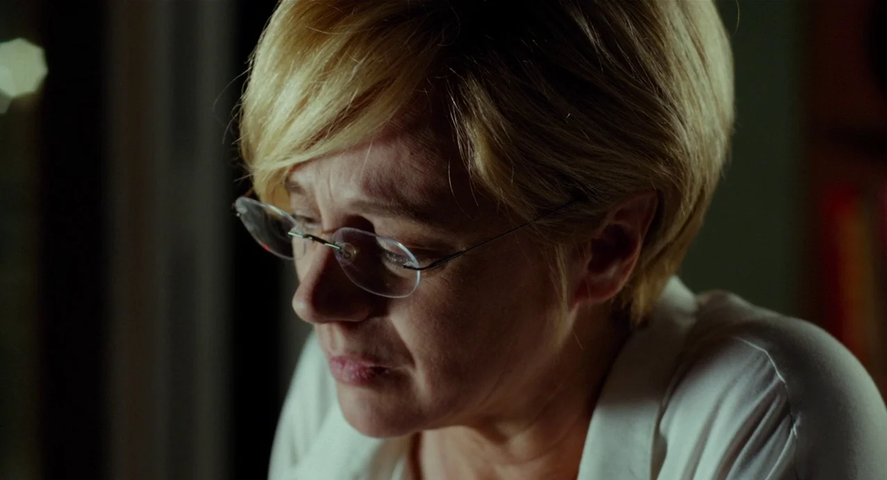 Julieta (2016) movie still: closeup — A close view of a woman indoors in a dimly lit setting. The woman wears wire-rimmed glasses and looks down…