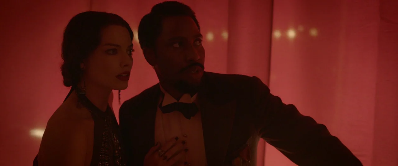 Amsterdam (2022) movie still: medium — A dark, enclosed space draped with fabric illuminated by a vibrant red light. A man in a tuxedo and a woman…