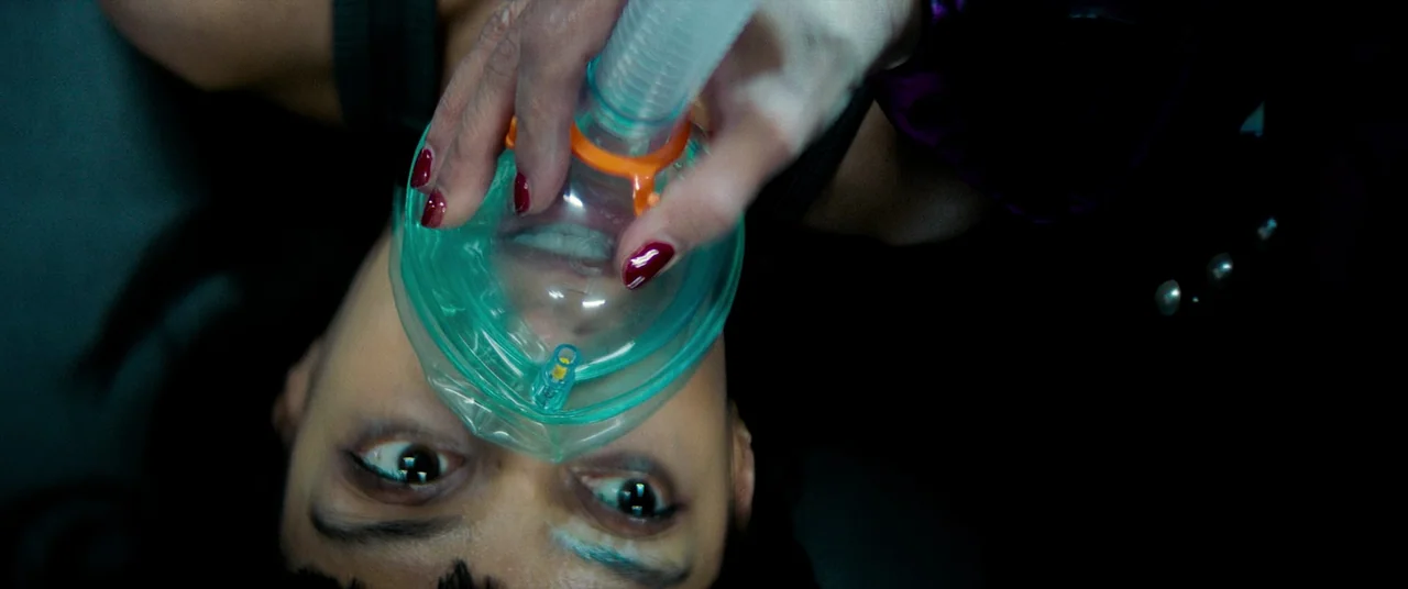 Polite Society (2023) movie still: closeup — A woman lies on a surface with an oxygen mask pressed over her face. The camera looks directly down at…